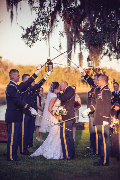 Bride and groom kiss under a military saber arch at an outdoor wedding.