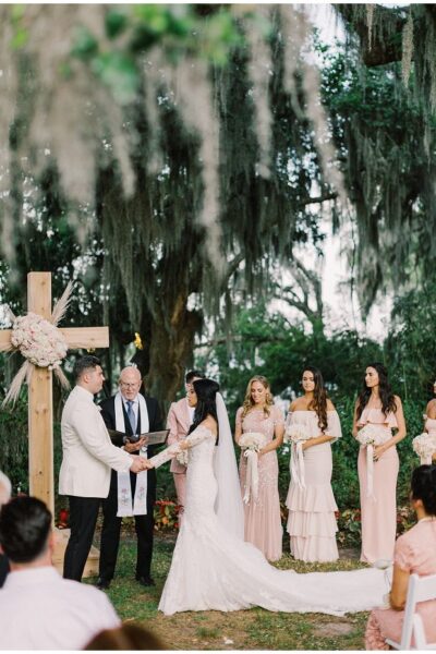 Bride and groom exchange vows outdoors surrounded by bridesmaids and guests.