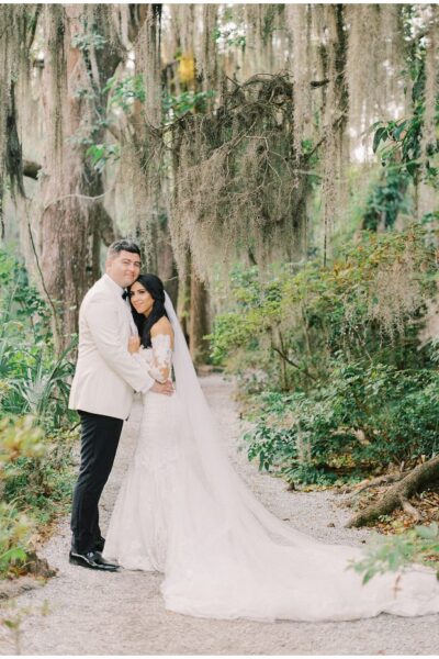 A couple embraces in a lush forest, the bride in a long white gown and the groom in a light jacket.
