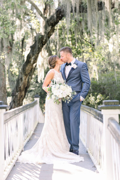 Newlywed couple shares a kiss on a scenic bridge surrounded by lush greenery.