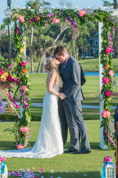 Bride and groom share a kiss under a floral arch during their outdoor wedding.