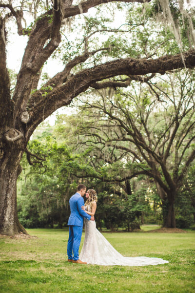 A couple embraces under large, arching trees in a serene outdoor setting.