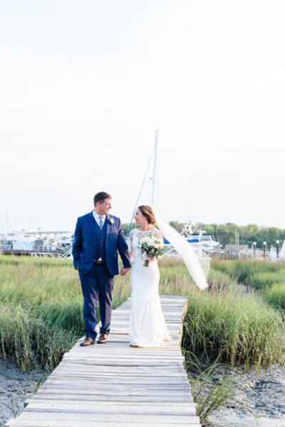 A bride and groom holding hands and walking near a marina on their wedding day.