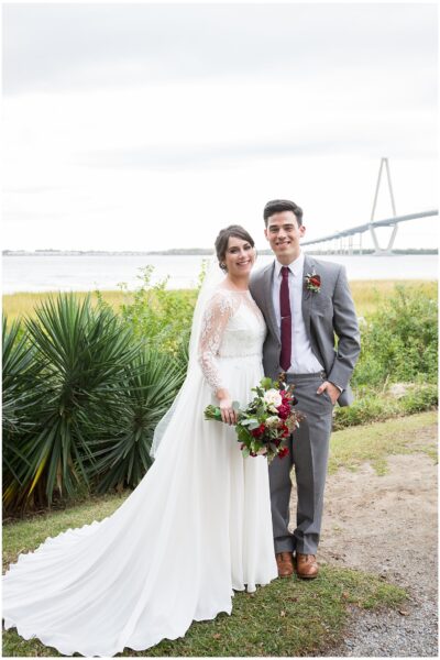 Newlyweds pose happily outdoors near a waterfront with greenery and a bridge in the background.