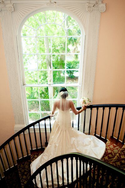 Bride in elegant gown standing by a large window with a bouquet.