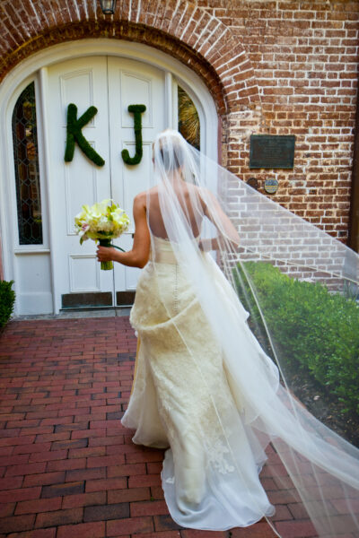 Bride in elegant gown walks toward building entrance with bouquet.