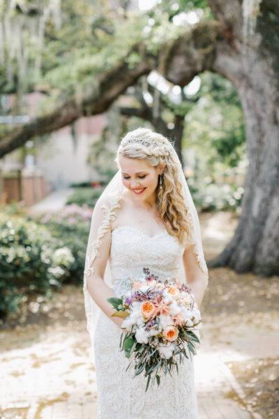 A bride in a lace wedding dress holding a colorful bouquet outdoors.