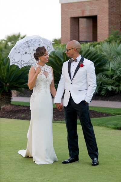 Bride and groom elegantly dressed outdoors, bride holding a parasol.
