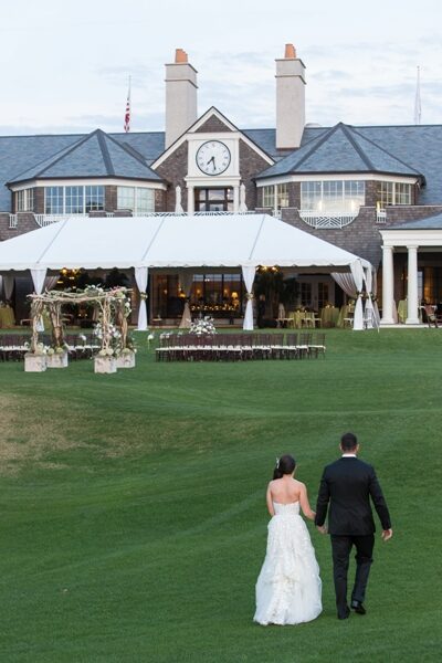 Newlyweds walk hand in hand across a manicured lawn towards a wedding reception tent.