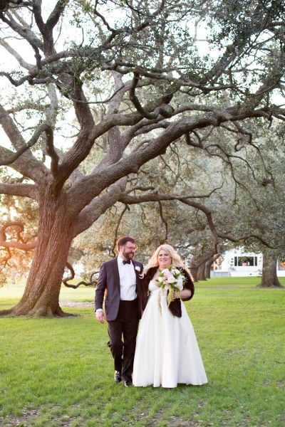Bride and groom walking under large oak trees at sunset.