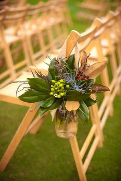 A rustic floral arrangement in a jar hanging on a wooden chair.