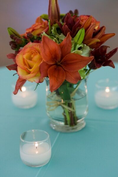 A bouquet of orange and yellow flowers in a clear vase with candles around.