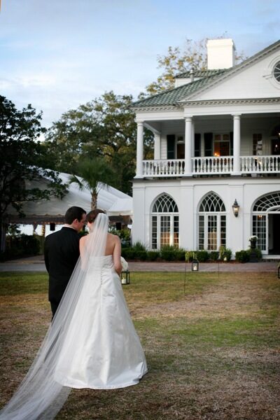 Newlyweds walk hand in hand outside a grand white mansion.
