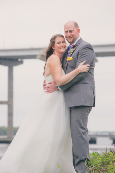 Bride and groom embracing outdoors with a bridge in the background.