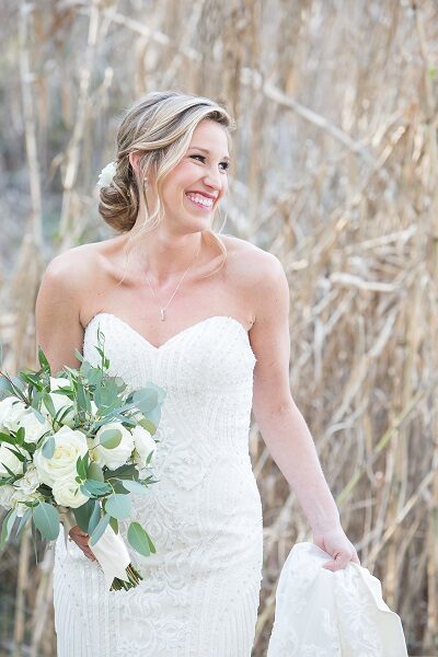 Smiling bride in a white strapless wedding gown holding a bouquet outdoors.