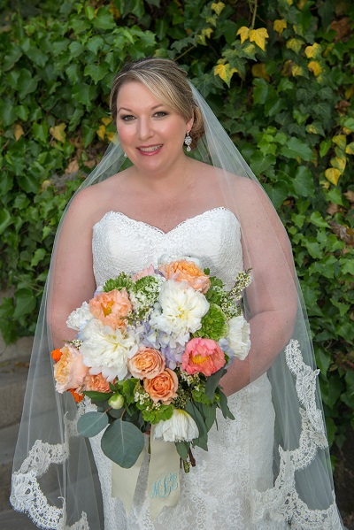 Bride in a white gown holding a colorful bouquet with a veil and greenery background.