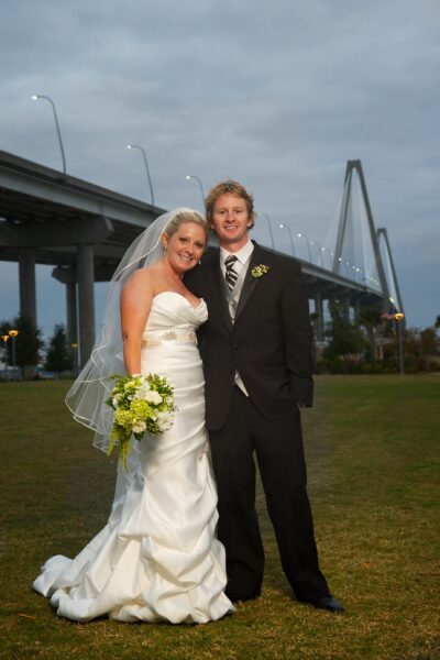 Newlywed couple posing outdoors near a bridge at sunset.