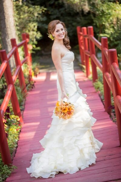 Bride in elegant gown holding bouquet on red bridge outdoors.