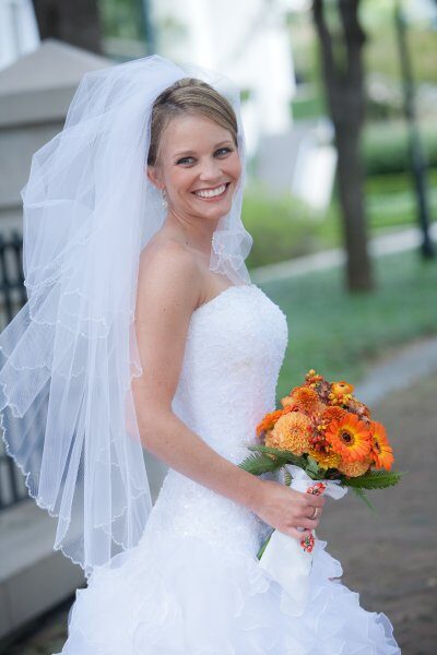 Smiling bride in white wedding dress holding a bouquet of orange flowers.