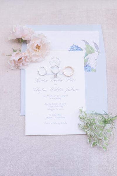 Elegant wedding invitation with floral accents and rings on soft blue background.