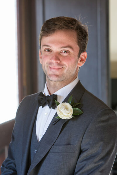 A groom in a dark suit smiling with a white rose boutonniere.