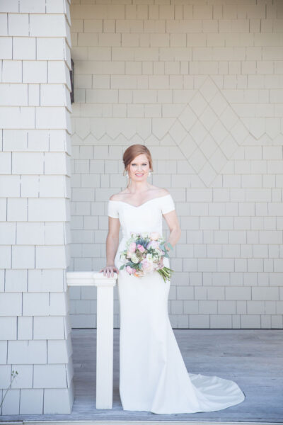 Bride in a white dress holding a bouquet, standing by a white bench against a light brick wall.