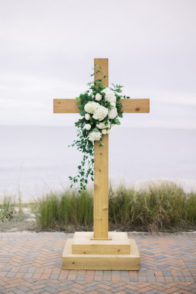 Wooden cross decorated with white flowers and greenery on a beach.