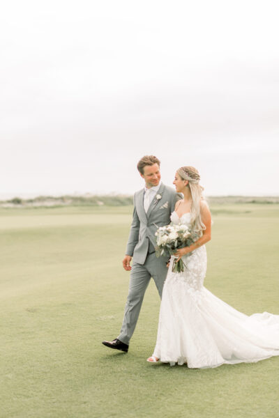 Bride and groom walking on grassy field.
