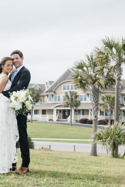 Newlywed couple embraces on a grassy lawn with beach houses behind them.