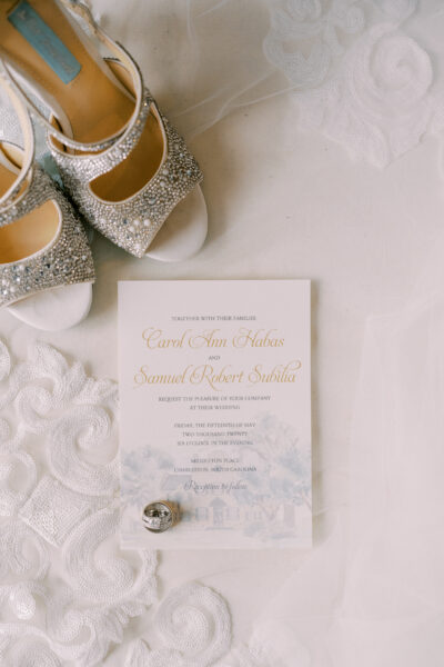 Elegant wedding invitation with silver shoes and rings on a lace fabric.