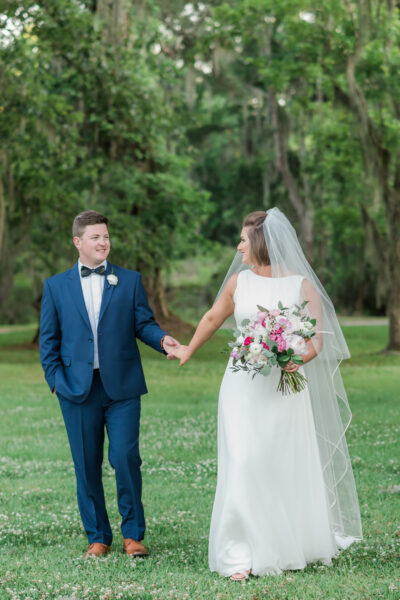 A bride and groom holding hands outdoors, smiling at each other.