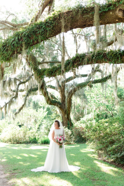 Bride in white gown holding bouquet under large mossy tree in a sunny garden.