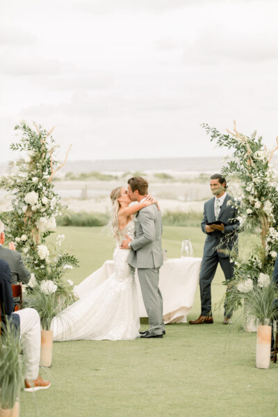 Bride and groom share a kiss at outdoor wedding ceremony by water.