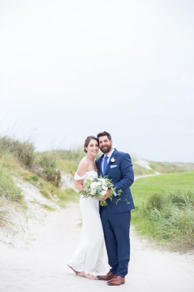 Bride and groom smiling on a sandy path by greenery on their wedding day.