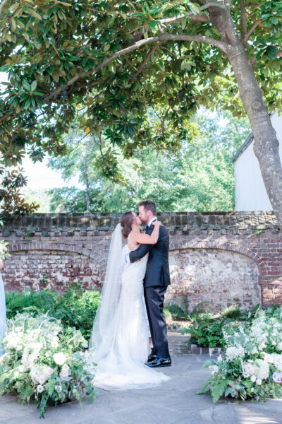 Bride and groom embrace outdoors by a brick wall under trees.