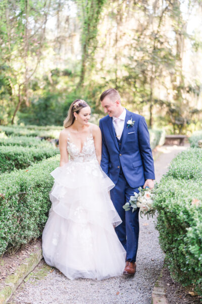Bride and groom walking hand in hand through garden hedges.