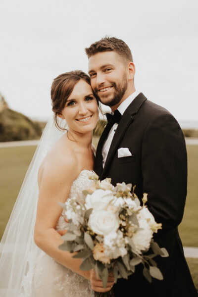 Smiling bride and groom posing outdoors, bride holding bouquet.