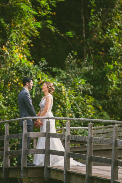 Bride and groom holding hands on a wooden bridge surrounded by green foliage.