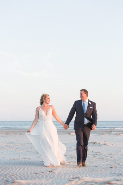 Bride and groom holding hands and walking on a beach at sunset.