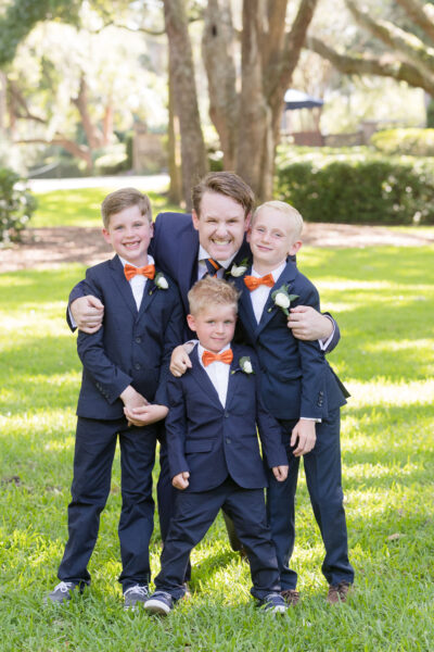 A man and three boys in suits smiling outdoors at a sunny park.