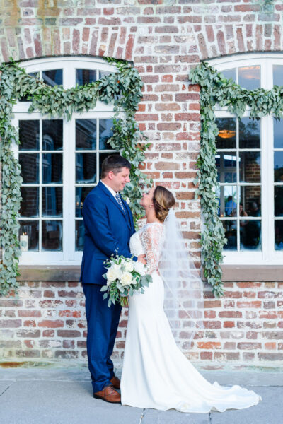 Bride and groom share a moment outside a brick building adorned with greenery.
