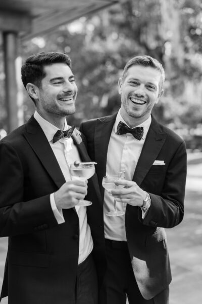 Two men in tuxedos smiling and holding cocktails at a formal event.
