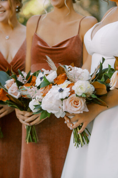 Bridesmaids in rust dresses and bride in white holding flower bouquets.
