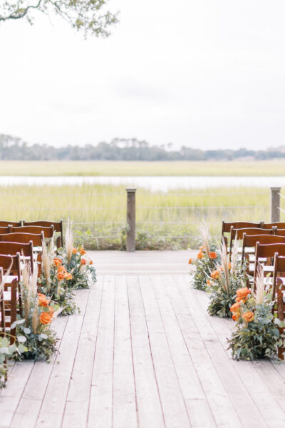 Outdoor wedding ceremony setup with flower-decorated wooden chairs facing a scenic field.