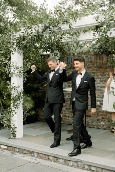 Two men in tuxedos joyfully walking outdoors at a wedding.