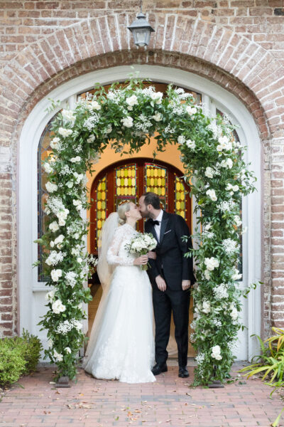 A newlywed couple shares a kiss under a floral arch at a brick venue.