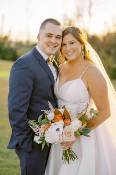 A happy couple in wedding attire smiling outdoors at sunset.