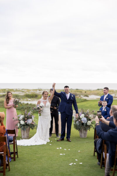 A joyful couple celebrates their wedding outdoors with guests applauding.