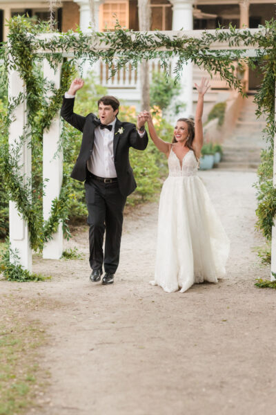 A joyful bride and groom celebrate outdoors with raised hands.