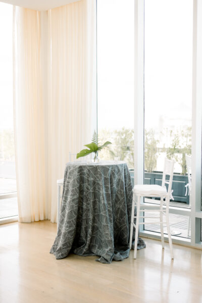 Round table with a patterned tablecloth and a small plant by a bright window.